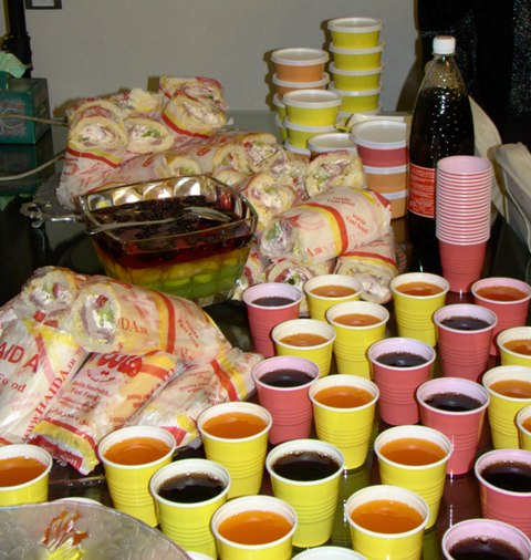 The image presents a colorful assortment of drinks and bites. There are numerous cups, bowls, and food items arranged on a table. The cups and bowls come in various sizes and colors, with some cups having drinks in them, ready to be served. The bowls appear to contain food, possibly wraps, which are in view. A dark-colored bottle is also present on the table, suggesting additional drink options. The array of items gives an impression of a buffet or party setting.