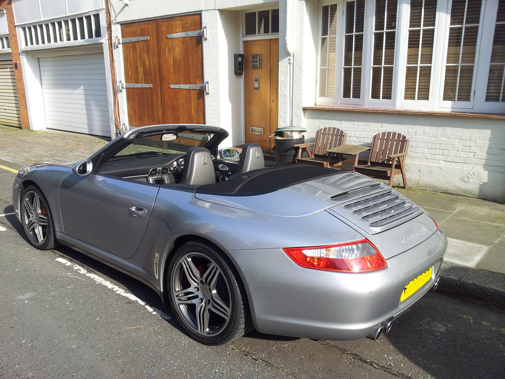 56 Plate 997.1 C4S Cab - Hold or Sell? - Page 1 - 911/Carrera GT - PistonHeads - This image presents a captivating scene of a gray Porsche 356 sports car parked on the side of a street. The car, featuring a black soft top and shiny silver rims, is parked in front of a building with a wooden door. It's positioned right next to a parking meter, indicating it's legally parked. The backdrop of the scene is an apartment building with beige walls and a black fence.