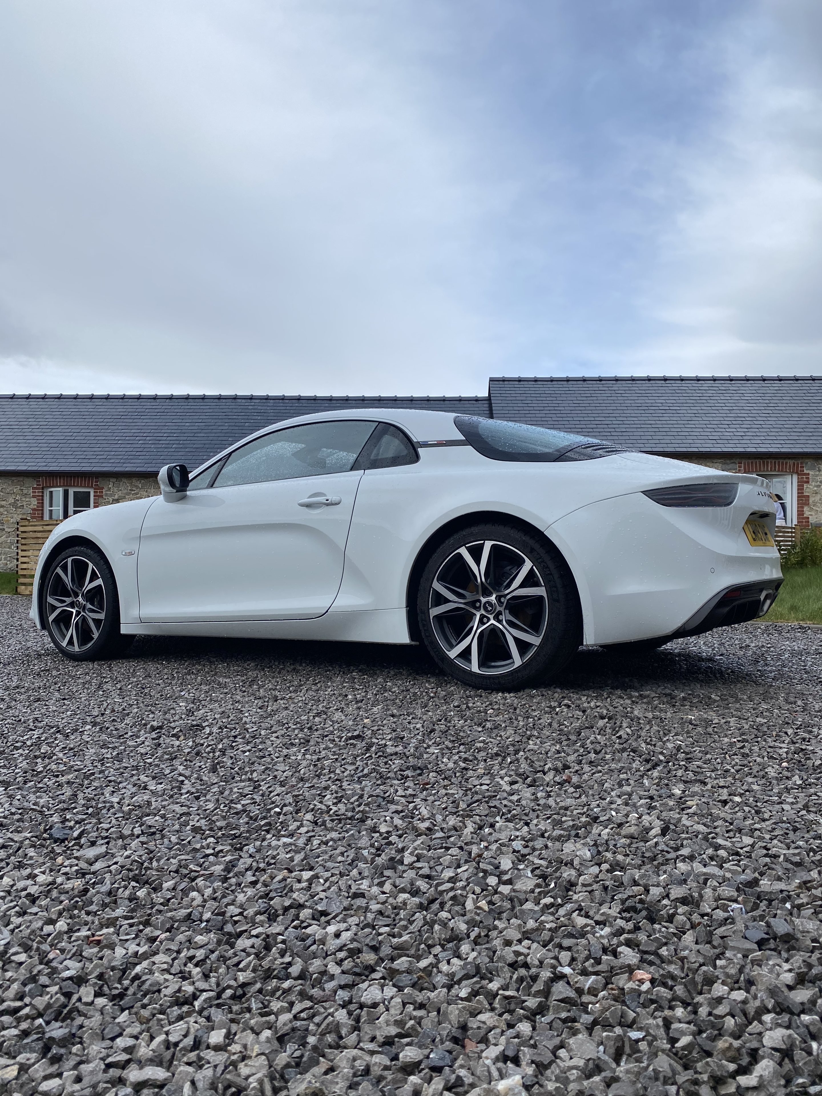 Alpine A110 owners - Page 48 - French Bred - PistonHeads UK - The image shows a white sports car parked on a gravel driveway. It's an open-top model, suggesting it might be used for convertible-style driving or is in the process of being converted. The car has a sleek design with visible aerodynamic features and alloy wheels. There's a license plate mounted at the back, but the text is not legible due to resolution. In the background, there is a building with a sloped roof, which appears to be a residential structure. The overall scene conveys a sense of luxury and exclusivity, often associated with high-end sports cars.