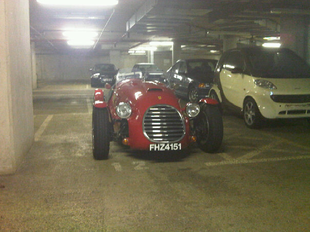 What is this? - Page 1 - General Gassing - PistonHeads - In the dimly lit parking garage, a vintage red sports car, complete with a convertible top, is parked. The car is positioned on a parking spot marked with a white line. It's situated between two other cars, with a yellow Chevrolet car visible in the background. The garage itself has a concrete floor and white walls, creating a stark contrast to the vibrant red of the sports car. The image captures the elegance of the vintage car amidst the mundane yet industrial setting of the parking garage.