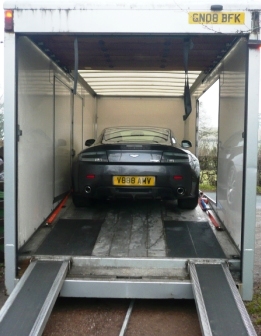 Photos attached - delivery day  - Page 1 - Aston Martin - PistonHeads - The image shows a car placed on a winch or lift system within the back of an enclosed trailer. The car appears to be modified as it has been lowered onto the lift platform, ready to be hauled away. The lift platform has rollers on each side, and there are several ratchets, suggesting a secure attachment. The trailer is surrounded by a high side, and the back wall is a solid structure, providing support during transportation. There are no visible texts or distinctive markings in the image.