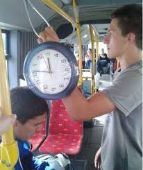 Big Watch's... - Page 1 - Watches - PistonHeads - The image shows an inside scene of public transportation, specifically a bus. A young man is seen holding a round analog clock. Another young man is looking up at the first, seemingly interested in the time displayed on the clock, which is just before noon, as indicated by the numbers 5 and 8 on its face. The clock is clear and readable with a red frame. In the background, multiple passengers fill the space of the bus, and the interior design suggests a typical transit setting.