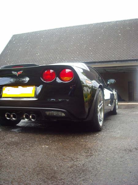 Clean Properly Pistonheads - The image portrays a sleek, black sports car parked in a garage. Its rear lights add a touch of color to the otherwise monochrome exterior. The car's license plate, reading "AB10", stands out against the dark background. The car appears to be parked on a rainy day, as evidenced by the wet floor in the garage. In the background, part of the car's side can be seen, further emphasizing the car's sleek and aerodynamic design. The image does not contain any text other than the license plate.