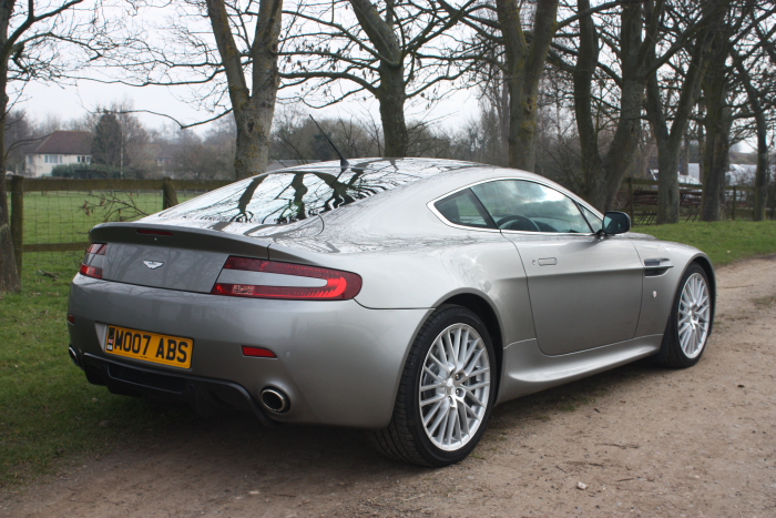 Difference Spot Pistonheads - The image showcases a silver Aston Martin sports car parked on a dirt road with trees in the background. The car is facing towards the right side of the photo. The car's vibrant red taillights contrast with its sleek silver exterior, and it is equipped with a spoiler, adding to its sporty aesthetic. The landscape and the setting suggest a serene and secluded location, away from the hustle and bustle of city life. The focus of the photograph is clearly the car, with its details of design and craftsmanship highlighted against the natural backdrop.