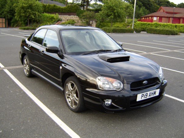 My Car - Page 1 - Readers' Cars - PistonHeads - The image features a black Subaru sports car parked in a lot with white striped lines. The car is positioned at a slight angle, facing towards the right side of the image. It has a black roof and black rims, giving it a sleek and sporty appearance. The license plate on the back of the car starts with the letters "PI9". The lot appears to be outdoors during daylight hours, with a paved surface and a red wooden building in the background.