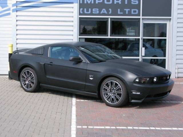 matt black 2011, not my cup of tea. - Page 1 - Mustangs - PistonHeads - The image shows a sleek black Ford Mustang with a bright sun glare, parked in front of a store. The car is positioned slightly to the left of the viewer's perspective, as if it has just pulled up to the building. The store behind the car features a prominent window display with the "USA J LTD" logo, suggesting that the store specializes in automotive imports. The overall scene gives off a vibe of travel or car shopping, with an emphasis on the flashy sports car.