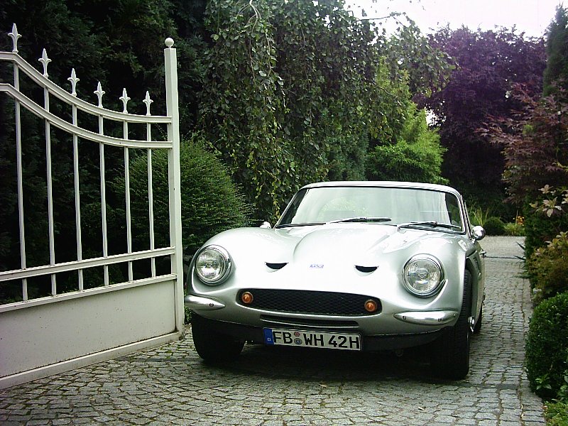 Early TVR Pictures - Page 64 - Classics - PistonHeads - The image illustrates a vintage silver convertible car parked in front of a white metal gate. The car, with its distinctive upswept headlights, is the focal point of the composition. The gate, positioned to the left of the car, contributes to the framing of the image. Beyond the gate, there is a verdant hedge, which adds a sense of greenery and serenity to the scene. The presence of the car and the gate suggests a sense of exclusivity and sophistication associated with the location.