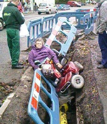 Pistonheads Extreme Cornering - The image shows a scene with multiple elements. In the foreground, there is a person sitting amidst what appears to be discarded items, including a dismantled motorcycle and a traffic barrier. The individual seems to be surrounded by various pieces of debris and material that could be the remains of a dig site or construction work. In the background, there's a greenish-blue obstruction in the middle of a road, possibly related to the situation in the foreground. A few cars and individuals can be seen further in the distance, suggesting that this scene takes place in a busy urban area.