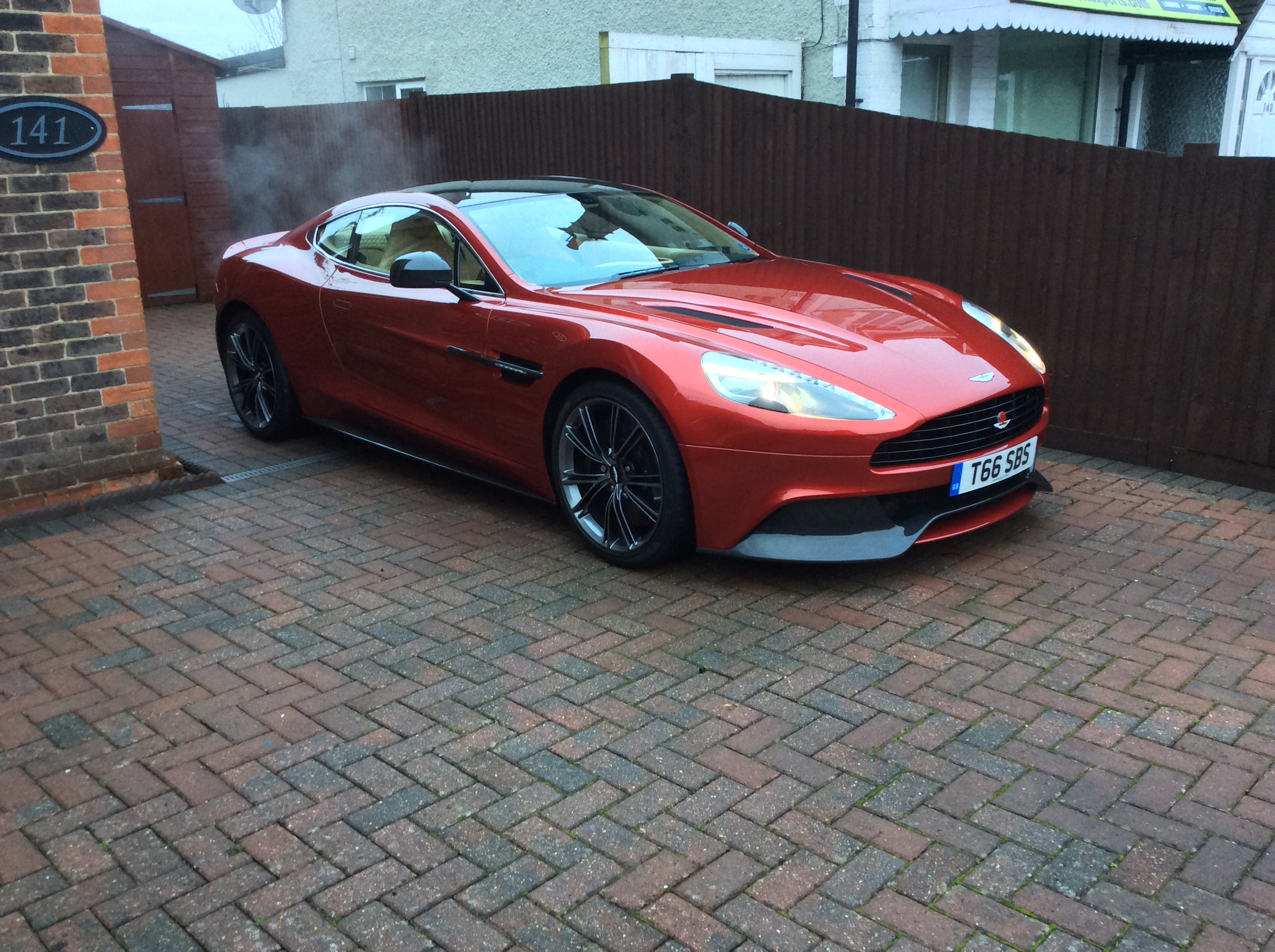 So what have you done with your Aston today? - Page 293 - Aston Martin - PistonHeads - The image presents a vibrant red Aston Martin sports car on a brick driveway. The car is parked diagonally, accentuating its sleek profile. On the right side of the frame, a fence made of vertical wooden slabs runs parallel to the car, adding a rustic contrast to the car's luxurious appearance. The Aston Martin bears a license plate reading "T66 BIN", and there are puddles on the ground, indicating recent rainfall.