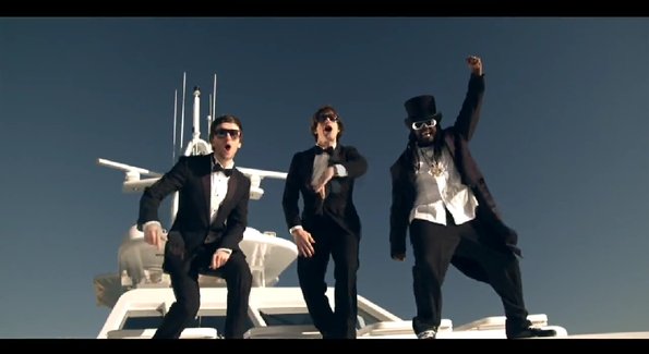 Boat Pistonheads - The image is a playful and dynamic scene. It features three men standing on the bow of a boat. Two of the men are dressed sharply in suits, complete with matching bow ties, dark sunglasses, and colored sneakers, conveying a hipster or possibly a parody of rappers' fashion. The third man stands out with his oversized white hat and a shirt with a furry collar, also in black. They pose with their hands up in the air, suggesting a celebration or a high feeling. The boat they are on is white and looks ready for an outing, indicating a casual, leisurely setting.