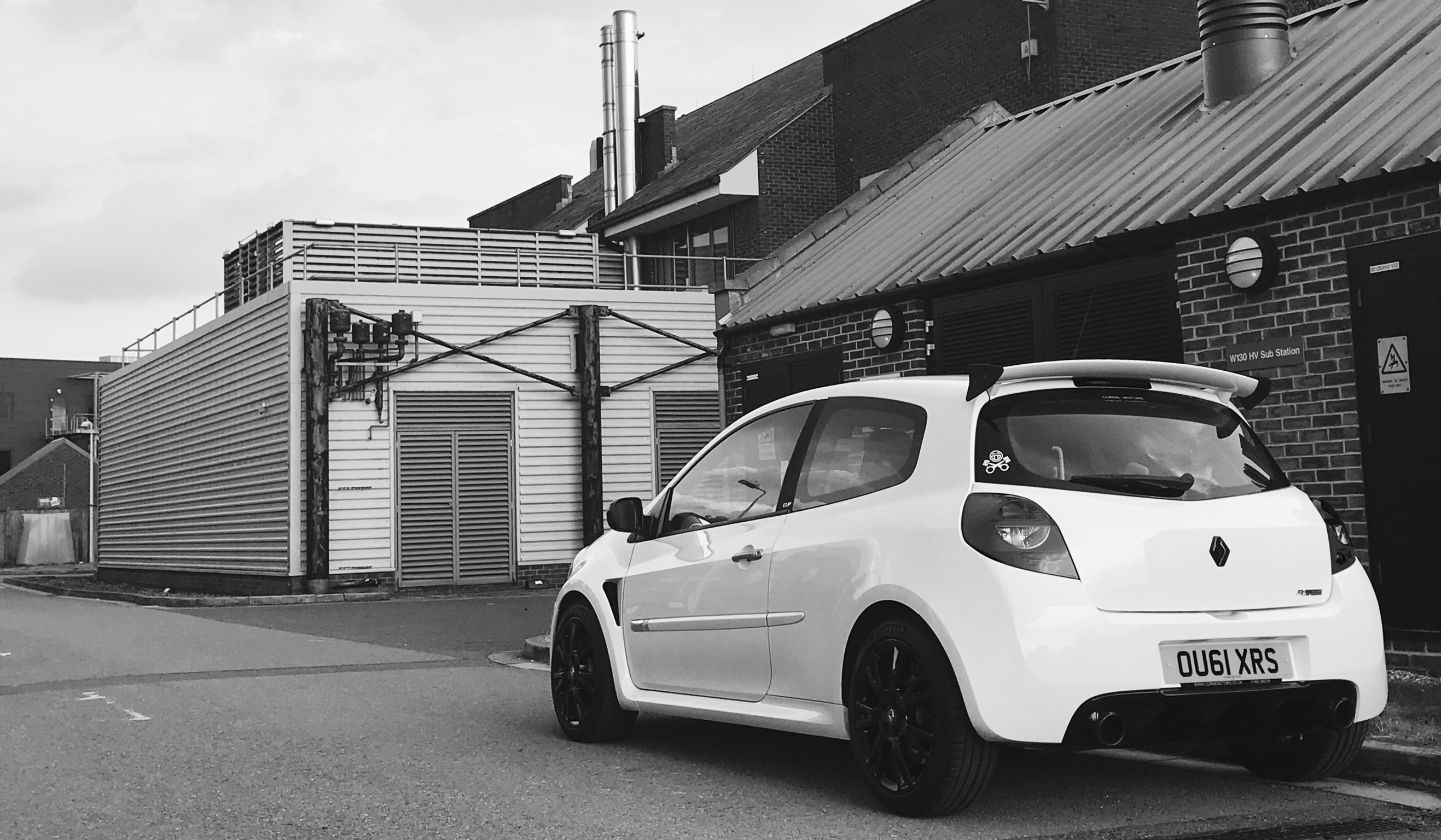 MrTouring car history and current e61 M5 and 944 - Page 3 - Readers' Cars - PistonHeads - This black-and-white photo features a vibrant little car parked on a street next to a building. The image appears illuminated with artificial lighting, possibly coming from nearby street lamps or artificial delivery dock lights. The vehicle is a hatchback with a distinctive front design, bearing the mark "DSUS 1KX" on its license plate. The surrounding buildings, partly obscured by the focus on the car, have a split-sided design with white panels and darker shades. Adjacent to the vehicle is a brick structure with a noticeable ventilation system on its roof.