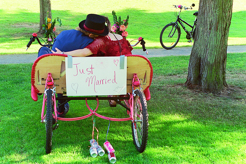 In the center of the image, a couple is dreamily seated under an umbrella in a charm carriage located in a park. The carriage is designed with heart-shaped accents, and it's adorned with flowers, conveying a romantic and festive atmosphere. On the background, a bicycle is parked, and a tree provides a pleasant shade. The scene is tranquil and evokes a sense of celebration and love.