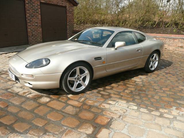 Your DB7 pictures. - Page 1 - Aston Martin - PistonHeads - In the image, a silver sports car is parked amidst a backdrop of a brick sidewalk and a glimpse of a red brick building. The car is positioned in such a way that it takes up a large portion of the frame. The sun shines on the car, reflecting off its sleek silver surface. The car's design, paired with the brick paving, suggests a neatly maintained urban or suburban setting. The focus of the image is clearly the car, drawing attention to its shiny exterior and impressive build.