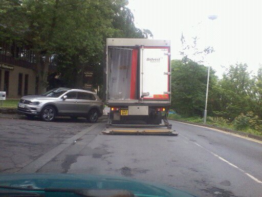 The BAD PARKING thread [vol3] - Page 344 - General Gassing - PistonHeads - The image depicts a street scene where a large, red and white truck is stopped on the side of the road at a crosswalk. The truck has a section of its trailer doors open, revealing some cargo inside. To the left of the truck, there is a silver car parked at the curb, presumably waiting for the large truck to pass to continue. The truck appears to be a standard delivery vehicle, with a license plate that reads "YE16IN". The overall setting could be an urban area with trees and grass visible in the vicinity, indicating a well-kept roadside. The lighting suggests it's daytime.