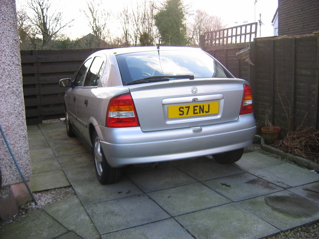 Rubbish Spotted Chav Plates Pistonheads - This image features a gray Honda Integra parked in a driveway. The car is positioned facing left with its front end visible. The Integra has a plate that reads 'S7 ENJ', which suggests it's registered in England. There's a wooden fence to the right of the car, and some verdant shrubs are also visible in the vicinity, providing a sense of a well-maintained garden. The photograph is taken during daylight under clear, blue skies, illuminating the calm, suburban setting.