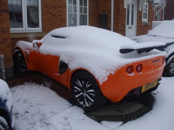 Exige Pistonheads Mini - The image captures a single, vibrant red sports car parked on the side of a residential street. It appears well-maintained and is uniquely characterized by a long, black spoiler attached to its rear wing. Coincidentally, the car is covered in a fresh layer of snow, making it stand out against the clear sky in the background. The street itself is bordered by buildings that, like the snow on the cars, add a sense of depth and tranquility to the scene.