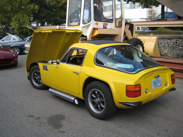 Early TVR Pictures - Page 36 - Classics - PistonHeads - The image depicts a vibrant yellow sports car with its hood raised, displaying what appears to be a heart-shaped cutout near the engine area. The vehicle is parked outdoors, seemingly at an event or exhibition involving vintage and unique vehicles. In the background, the scene features an orange car, several trucks, a fence, and trees, creating a colorful and outdoor setting. Additionally, there is construction equipment visible in the background, suggesting that the event might be taking place in a location that is being repaired or renovated.