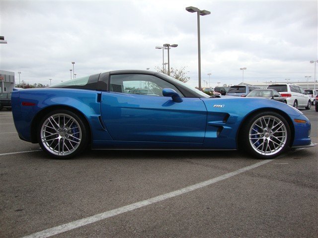 I finally gave in....... - Page 1 - Corvettes - PistonHeads - The image captures a striking blue Corvette, a sports car known for its performance and style, parked on a paved parking lot. The car is positioned centrally in the frame, drawing the eye with its vibrant color and sleek design. Its wheels are silver, adding a touch of contrast to the overall blue tone. The setting appears to be a public parking area, with a few other cars visible in the background.