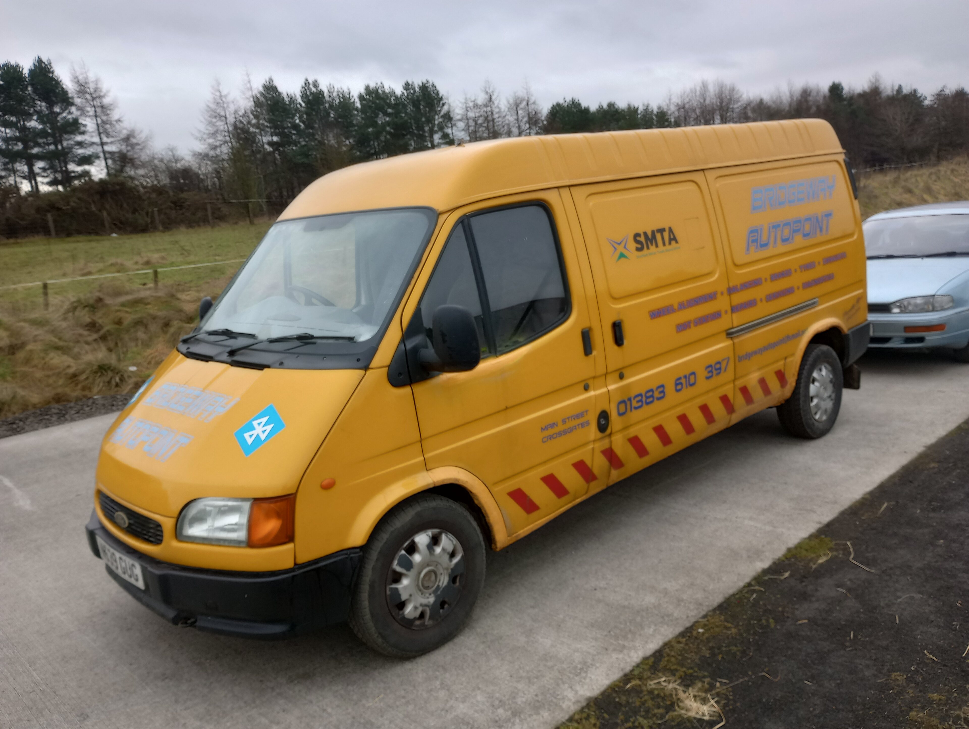 The Art of Shedding  - Page 32 - Readers' Cars - PistonHeads UK - This image features a van parked on what appears to be a rural or semi-rural area. The van is predominantly yellow and has a sign in the front window that says "S&C" with a phone number underneath, suggesting it may be used for commercial purposes such as transporting goods. There's also a noticeable sign with more information on it, but it's not entirely clear from this image. The van is parked on what seems to be an asphalt surface near a field or pasture. The sky overhead is cloudy, and the lighting in the photo indicates either dawn or dusk. There are no people visible in the image, giving a sense of quietness to the scene.