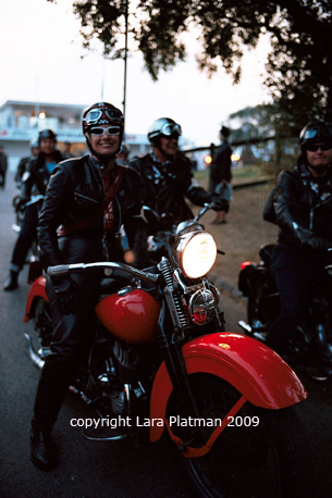 2009 Revival pictures - Page 6 - Goodwood Events - PistonHeads - The image depicts a group of motorcyclists on a road, with a person seated on a red motorcycle in the foreground. The motorcyclist is wearing a helmet and sunglasses, and is looking to the side. The background is slightly blurred, but shows other motorcyclists on the road, creating a sense of motion and camaraderie. The image has a watermark text at the bottom that reads "Copyright Lara Platman 2009."