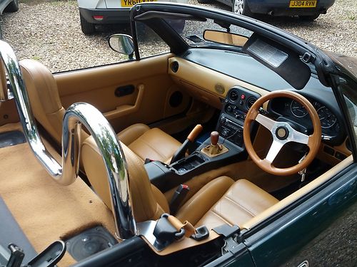 1991 mx5 limited edition- Rare i think pls help? - Page 1 - Mazda MX5/Eunos/Miata - PistonHeads - The image showcases an interior view of a vintage car with a beige leather interior. The steering wheel is prominent in the top right corner. The door is open, revealing the driver's seat inside. Outside, several cars and airplanes can be seen, indicating a busy or crowded space. The vintage car appears to be parked or stationary given its orientation and the surrounding vehicles.