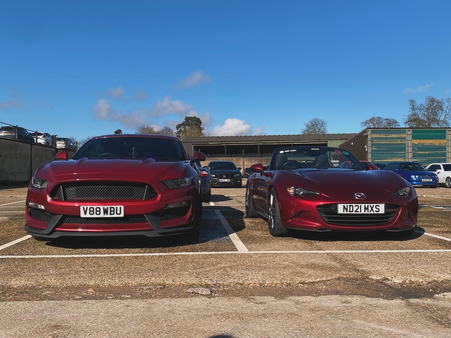 Pistonheads - The image shows two parked cars in a parking lot. The car on the left is a vibrant red, and it appears to be a modern sedan model. On the right, there's another car, which seems to be an older or classic model with a different design aesthetic. In the background, there are buildings and what looks like a commercial area with various signs. The sky above is clear, suggesting it might be a sunny day. There are no people visible in the image.