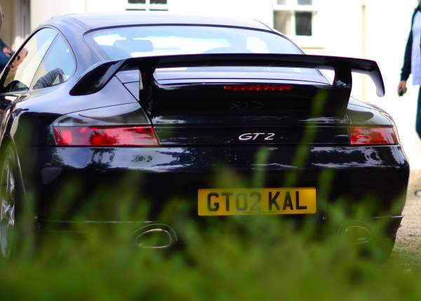 The image showcases the back view of a sleek black sports car. The car bears a yellow license plate that reads "GT02KAL". The license plate is set against the backdrop of lush greenery. The car's visible taillight and spoiler further accentuate its sporty appeal. In the background, there is a person partially visible, suggesting that the photo may have been taken from the side of a road.