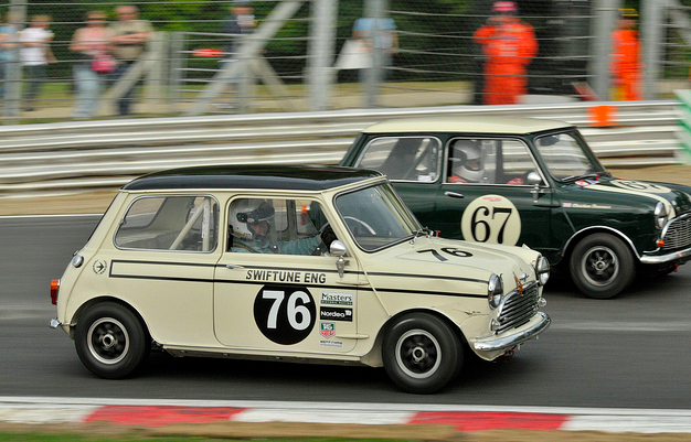 Just bought my self a Mini! - Page 1 - Classic Minis - PistonHeads - The image captures an exciting moment in a vintage race car event. Two iconic small race cars are in motion on a race track. The car on the left is number 76 and is white with a black stripe. The car on the right, number 67, is green with a white stripe. Both cars feature the number 76 prominently on their front. In addition to the two race cars, other elements like people and a fence are visible in the background, adding to the atmosphere of the event.