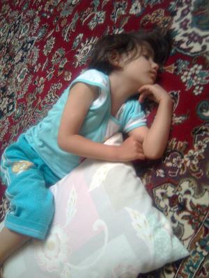 This image features a young child, most likely a girl, lying on her side on a surface with a geometric pattern, possibly a carpet. She is wearing a light blue t-shirt and blue pants, and there is a white pillow to the right of her head. The child appears to be asleep or very relaxed, with her forehead resting on her hand and her curly hair fanned out across the surface. The scene is serene, capturing a moment of rest and perhaps comfort.