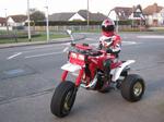 Pistonheads Illegal Thought - The image shows a young person riding a quad bike on a paved road. They are dressed in safety gear, including a helmet, which is red and white. The quad bike appears to be a red and white machine with a robust design, featuring fat wheels and a tubular frame. The person is positioned in the center of the frame, slightly leaning forward, indicative of movement. The background includes houses and grass on either side of the road, suggesting a residential area.