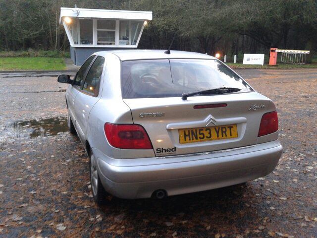 My Two French beauties. - Page 2 - French Bred - PistonHeads - In the image, a silver hatchback car, adorned with a black "Shed" sticker on the back, is parked on a wet asphalt surface. The car is positioned next to a lamp post that stands out against the dark sky, suggesting it's an overcast day. The car's license plate is clearly visible, reading "HNS53 YRT". The overall scene conveys a quiet, possibly outdoor, setting during the evening.