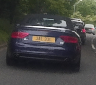 A car is parked on the side of the road - Pistonheads - The image shows a dark-colored sedan, likely a model of Jaguar, driving on a busy road. The vehicle's license plate is yellow and appears to be digitally obscured. The rear view image suggests a traffic camera or a rear camera of another vehicle that is capturing the movement of the Jaguar. The road itself is flanked by trees and buildings typical of an urban or suburban setting. The sky is overcast suggesting a completely cloudy, possibly gloomy day.