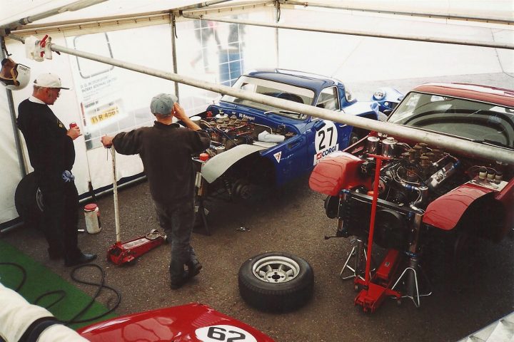 Early TVR Pictures - Page 28 - Classics - PistonHeads - The image captures a bustling scene in an indoor workshop where a group of people are working on vintage cars. There are three main cars in the scene, each mounted on a hoist. The cars themselves appear to be undergoing some sort of maintenance or restoration work. In the foreground, two individuals are engrossed in their tasks, one of whom is holding a drill, while the other is focused on a charcoal can, possibly for cleaning or fire suppression. The workshop is well-equipped, with tires scattered about and tools within easy reach. The overall atmosphere is one of intense focus and shared passion for automotive restorations.