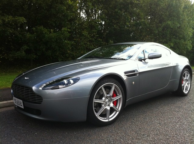 Pistonheads - The image features a sports car parked on the pavement alongside a road. The vehicle is a silver and black Aston Martin. It has a sleek design, characterized by a long front hood and a sloping rear windshield. The car is positioned at an angle that allows a clear view of its front and part of its left side. The Aston Martin logo is prominently visible on the front grille. The setting appears to be a sunny day as indicated by the shadows cast by the trees in the background. The car is set against a scenic backdrop of greenery and trees, suggesting a calm and sunny day.