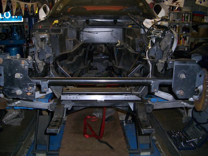 Pistonheads Rebuild Viper - The image shows an open vehicle's body shell, viewed from an angle looking downward from above. The car's structure is exposed, revealing the complexity and design of the vehicle's chassis, which includes various metal components and fasteners. Inside, the frame of the vehicle is visible, with black suspension parts, such as the bumper and components attached to it. The surroundings of the vehicle suggest that it is in a workshop or garage, evidenced by tools, equipment, and mechanical parts typically found in such environments.