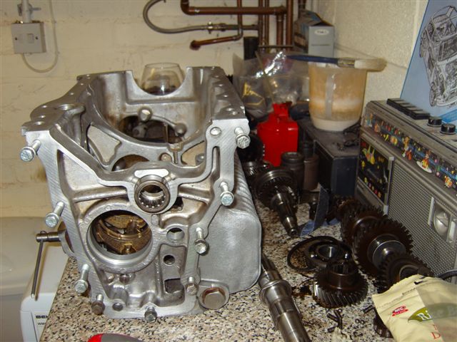 pics - Page 1 - Classic Minis - PistonHeads - The image shows a mechanic's workspace in a garage. There's a shiny silver engine block prominently placed on a workbench. Several automobile gears are scattered on the table, some chrome and others likely aluminum or steel. In the background, there is a typical garage setting with a shelf that has a collection of tools and spare parts. There's also a red cooler, a power drill, and a water radiator, adding to the industrial feel of the scene. The overall tone of the image suggests a professional environment dedicated to vehicle repair and maintenance.