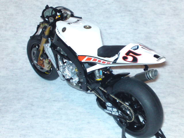 Yamaha Tamiya Pistonheads Moto - The image features a motorcycle with a predominantly white and black color scheme. It is a limited edition model symbolized by the number "5" on the front. The motorcycle is positioned at a slight angle, showcasing its profile, and the background is a plain, light surface, likely a surface they can detach to duplicate the models. A small detail, like a chain, is also visible, suggesting functional components of the motorcycle. The overall style of the image is realistic and detailed, providing a clear view of the motorcycle's design and features.