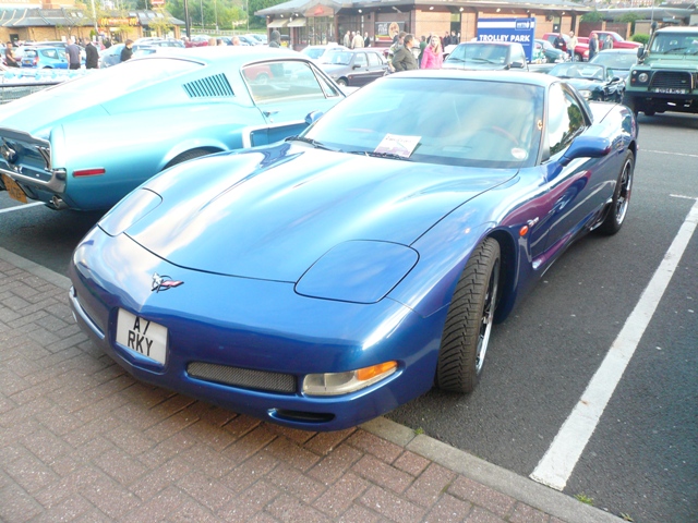 Pistonheads Metrocentre - The image captures a vibrant scene in a parking lot. Dominating the foreground is a striking blue sports car, its polished finish contrasting with the rough texture of the tarmac. The car is parked amidst an array of other vehicles, creating a dynamic tableau that fills the frame.

In addition to the sports car, there are several other vehicles in the lot, including noteworthy classics from the 1960s. These cars add a touch of nostalgia to the modern sports car, suggesting a fusion of past and present in this car show.

The image is shot from a low angle, offering an upward perspective of the cars. This angle places the sports car in the center, drawing the viewer's attention to it first before leading the eye across the other vehicles and architecture in the background.

Overall, the image is a lively snapshot of a car show, filled with colors, shapes, and textures that celebrate the rich history and diverse styles of automobiles.