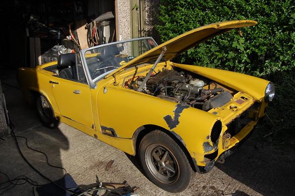 Healey Pistonheads Austin Sprite - This image shows a yellow classic car with the hood open, revealing a complex engine and various mechanical parts. The car appears to be in a process of restoration or repair, as indicated by the exposed engine, and the setting suggests a garage or workshop environment. The overall condition of the vehicle suggests it has undergone significant use or age, perhaps as a vintage, fun addition to a collection or for use in a specific event or project.