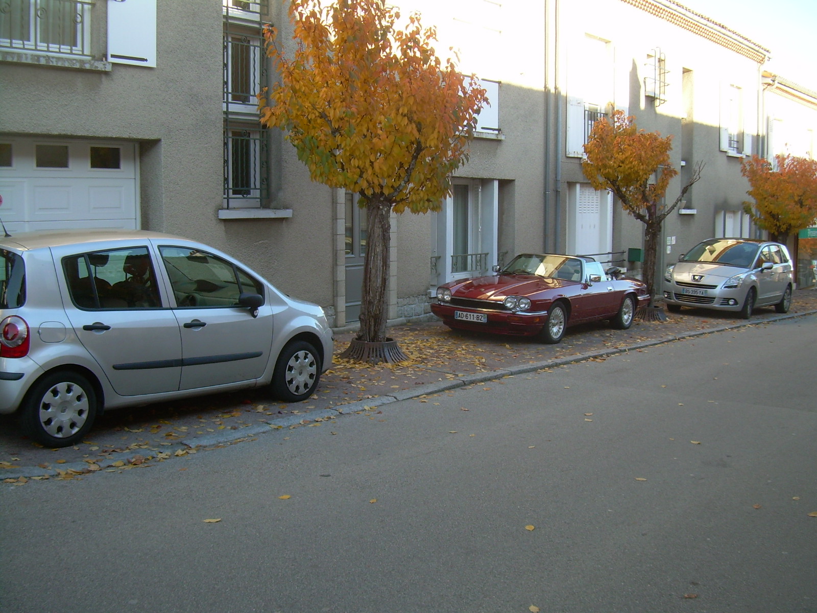 COOL CLASSIC CAR SPOTTERS POST!!! - Page 325 - Classic Cars and Yesterday's Heroes - PistonHeads - The image presents a serene scene on a residential street. Dominating the foreground are gray cars, parked neatly along the left side. The majority of the image is occupied by a gray car, which takes up most of the space. An older red car is parked further down the road, providing a vibrant contrast to the gray cars. The street itself appears to be in an urban location, with stone buildings siding the road. The trees and leaves dotting the background add a touch of nature to the urban setting. The arrangement of the cars and the overall atmosphere suggest a quiet morning or afternoon in this neighborhood.