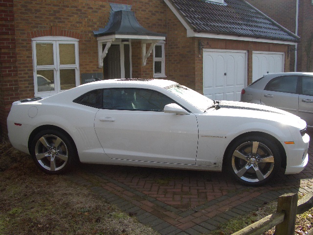 2010 Camaro - Page 1 - Yank Motors - PistonHeads - The image features a white two-door sports car parked outside a brick house. The car is positioned perpendicular to the curb, with one side turned to the left. It has a sleek design, characterized by a sloped roofline and a profile featuring a sharp edge and a lightly contoured bonnet. The house's architecture includes a brick facade with a contrasting half-timbered section. A small white canopy is attached to the brickwork above the front door, offering shelter. In the foreground, there is a wooden fence to the left, and part of another vehicle can be seen on the right.