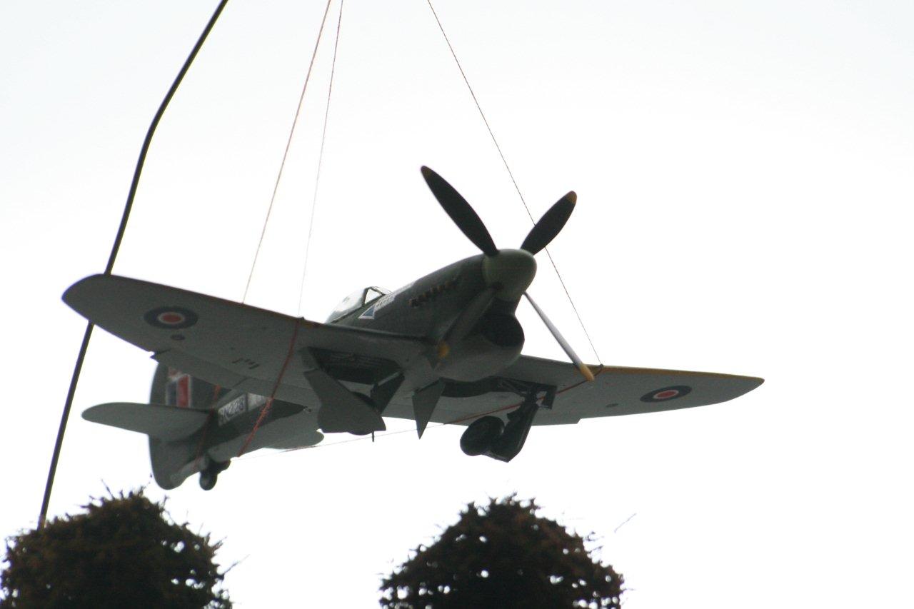 Academy Tempest Pistonheads Mcdjl - The image shows a vintage military plane hanging from a crane as it is being transported or raised in the air. The plane has a propeller, indicative of its early era design. The clear sky forms the backdrop, high above two distant trees at the lower edge of the frame. The perspective of the image gives the impression that the plane is suspended mid-air. The plane itself appears to be a historical military aircraft.