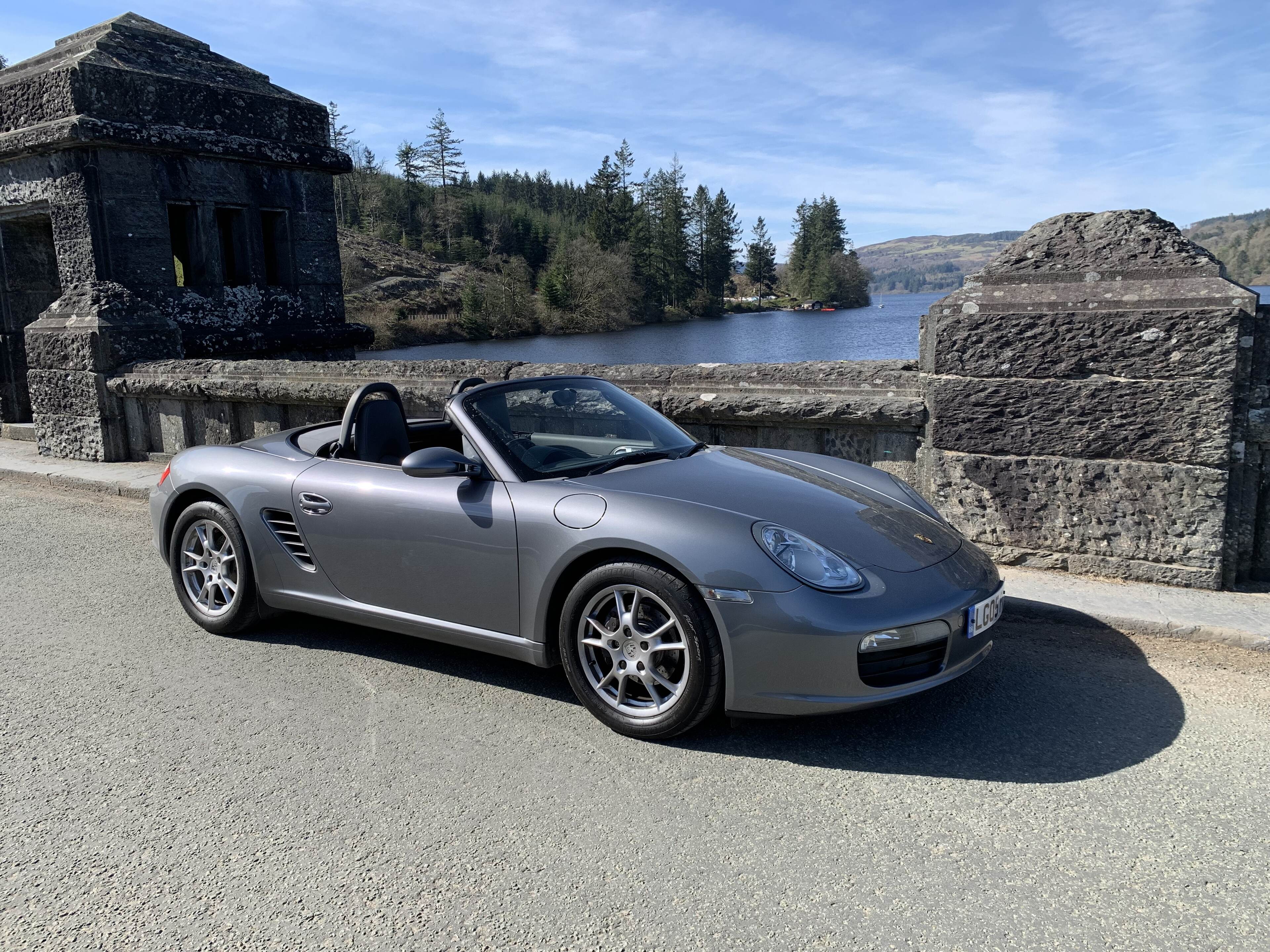 Pistonheads - The image shows a convertible sports car parked by the side of a road. The car has a sleek design with a silver color and the top is down, allowing us to see the interior. It's positioned in front of a stone structure that could be part of an old building or bridge. The environment suggests it might be near a lake or a river, given the presence of water in the background. There are no people visible in the image.