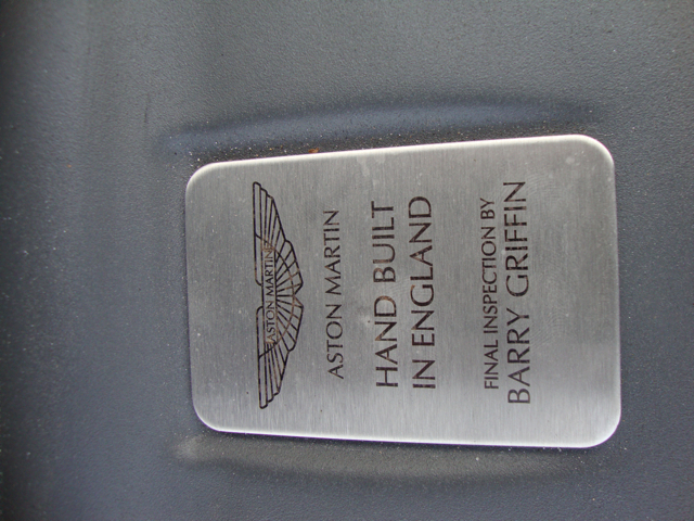 NAME PLATES - Page 1 - Aston Martin - PistonHeads - The image features a close-up of a metal sign or badge with inscriptions on it. The sign is silver with black lettering. The most prominent line of text reads "ASTON MARTIN HAND BUILT IN ENGLAND." Below this, there are two names providing commissioning information: "Griffin," which suggests the name of the commissioning agent or dealership, and "Finish," which might indicate the owner's origin or nationality. The text is arranged on multiple lines, with "ASTON MARTIN" and "HAND BUILT IN ENGLAND" occupying significantly more space than the names below. The overall look of the sign is sleek and professional, typical of vehicle brand identification.