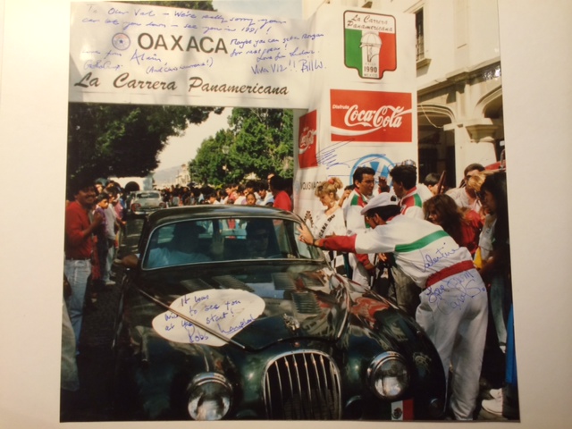 RE: Classic Jaguar unlocks Pink Floyd past - Page 2 - General Gassing - PistonHeads - The image is a photograph featuring an automobile in the center surrounded by several people. On the front of the vehicle, there's a classic car cartoon advertisement with a Coca-Cola logo in the upper part. The text on the vehicle includes "OAXACA" and names that appear to be signatures. The scene takes place outdoors with a crowd of individuals in the background, suggesting a special event or gathering. The style of the car and the billboard indicate a possible vintage or nostalgic theme. The photograph has a soft, natural frame with a bluesignature matt boarder, giving it an elegant and vintage feel.