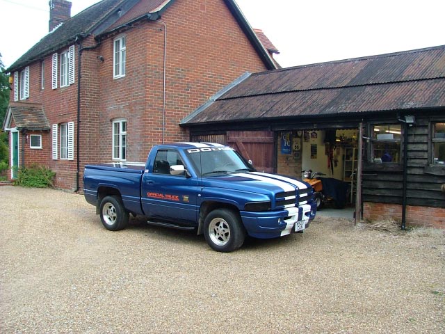 The image shows a blue Dodge Ram pickup truck parked in a gravel driveway. The truck has a vibrant blue paint job with white and silver stripes on both sides. It's an older model, with a visible V8 emblem on the passenger's side of the hood. Behind the truck, there is a classic red brick house with white window frames. The setting appears to be a residential or suburban area.