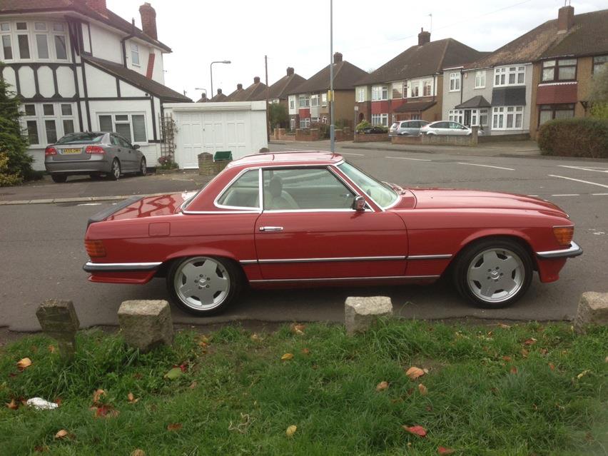 My 1979 Mercedes 450SL - Page 1 - Readers' Cars - PistonHeads - The image portrays a tranquil suburban street scene. At the center of the frame, a classic red sports car is parked, exuding a silent charm that contrasts with the everyday life. The car is sleek and glossy, parked on a grassy area, surrounded by neatly cut grass. The sky above is overcast, suggesting a calm and cloudy day.

In the background, quaint houses line the street, adding to the idyllic suburban setting. Their presence suggests a residential area, perhaps a quiet neighborhood. Further down the street, a few more cars can be seen parked, possibly belonging to the residents of the houses.

The road appears to be wide and well-maintained, suitable for regular traffic. Despite the mention of old cars, the image gives a general impression of a well-kept suburban area, where homes and cars are in harmony with each other. The absence of any people or movement in the image reinforces the serene, almost quiet atmosphere.