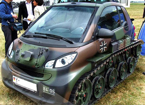 Pistonheads - The image shows a military-style car parked in a field. The car is green and black, with a camouflage design, and it features a for instance of the number 43. The vehicle has a compact design with visible tracks intended for off-road use. There seem to be small vehicles or components partially visible in the background, possibly part of the same military setup or event. There are people gathered around the car, suggesting it might be on display or part of a demonstration. The license plate and text read "84-005" in what appears to be a German military context.