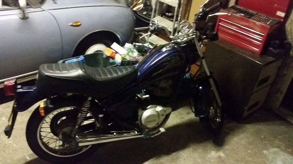 A motorcycle parked on the side of a road - Pistonheads - The image shows a parked blue motorcycle with a black seat and matching blue fenders. The motorcycle is leaning slightly to the left and is placed in what seems to be a garage. In the background, there are several objects, including another vehicle, a toolbox, and a red container. The foreground shows the front wheel of the motorcycle and part of the blue fender. There are no visible texts or distinct brands in the image.