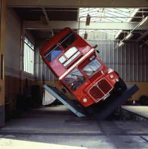 A red double decker bus driving down a street - Pistonheads - The image shows a captivating scene inside a workshop or warehouse-like setting. Dominating the frame is a large, red double-decker bus, tilted to the side on a ramp, defying the laws of gravity by seemingly hanging sideways. The presence of the bus itself, upside down and defying typical viewers' expectations. The ramp and the bus create a sense of depth in the image, while the workshop surroundings are characterized by a large window in the background, letting in natural light. The entire scene conveys a sense of curiosity and wonder about the circumstances of the bus's position and the possible alterations that were made to bring it to this unusual state.